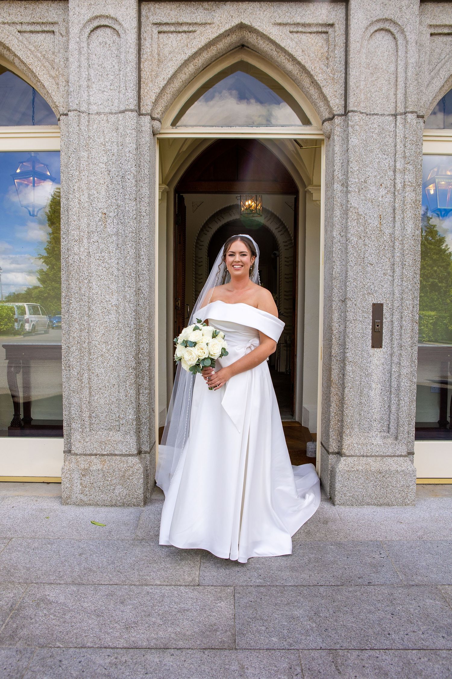 Bride in off-shoulder white gown holding bouquet stands in stone church doorway with veil and elegant train.