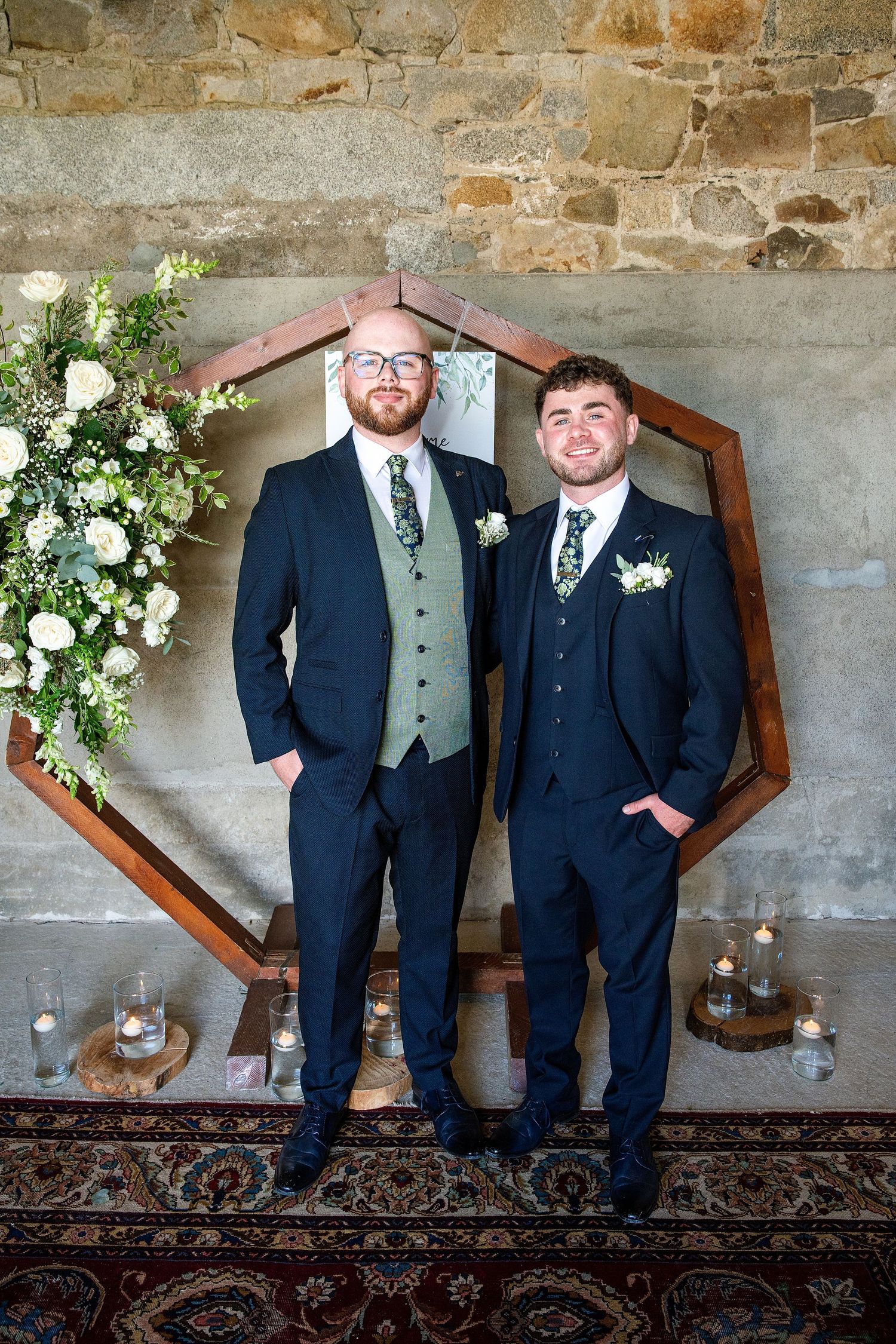 Two grooms in navy suits pose before a wooden hexagon arch adorned with white flowers at their wedding ceremony.