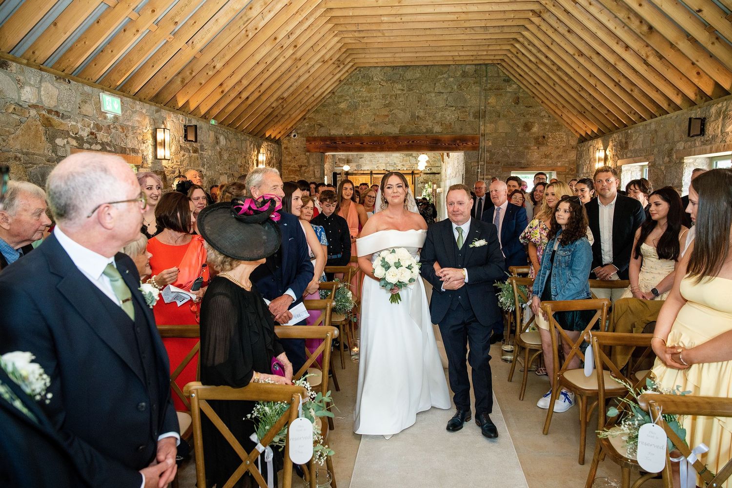 Bride walks down the aisle with escort in rustic stone barn wedding venue filled with seated guests.