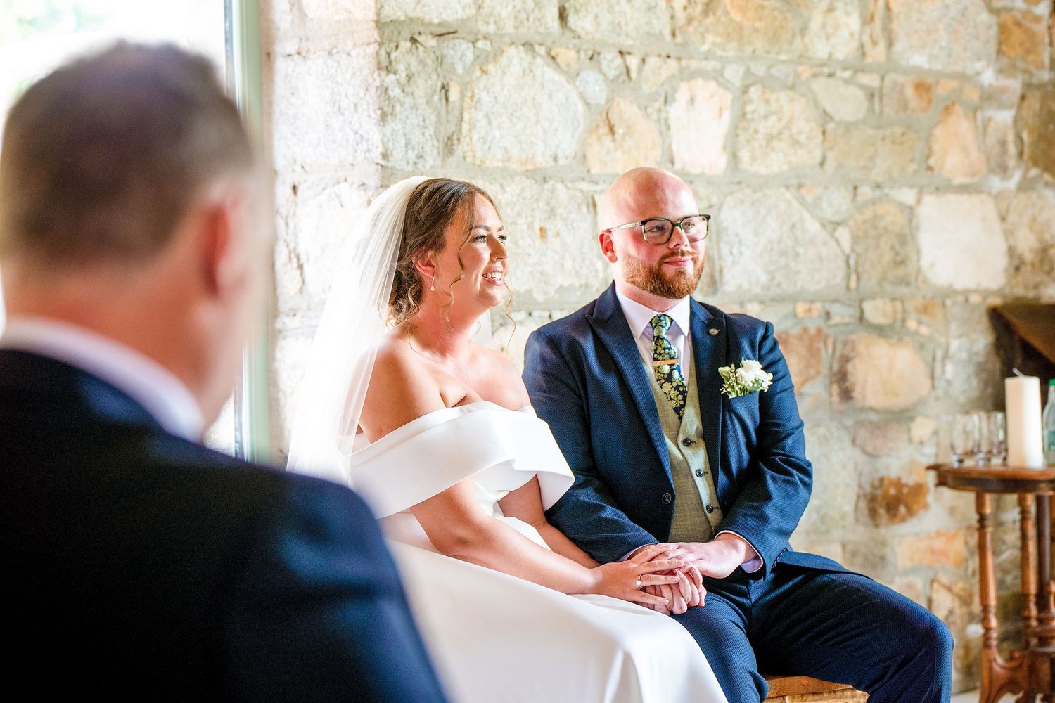 Bride and groom holding hands during wedding ceremony in rustic stone venue, bride in off-shoulder gown, groom in navy suit.