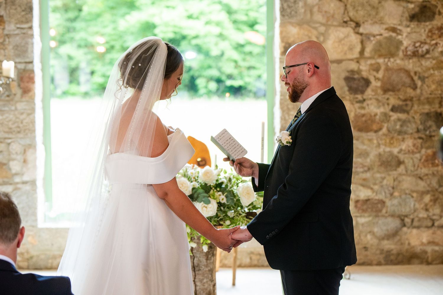 Bride and groom exchanging vows at a rustic stone wedding venue, bride holding white floral bouquet.