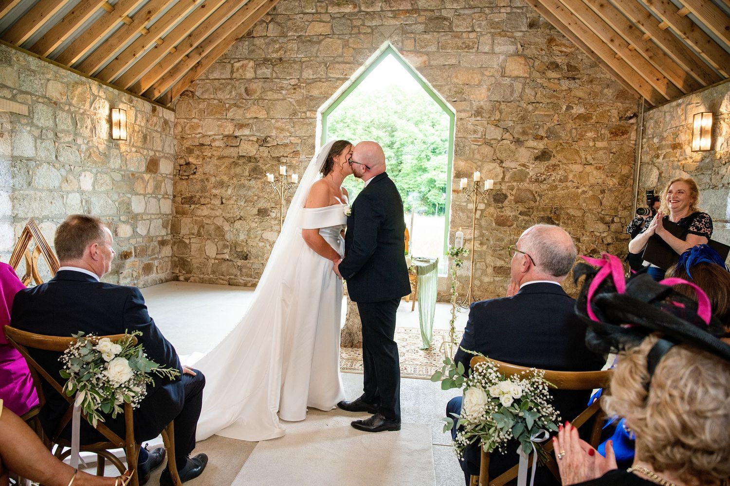 Bride and groom share first kiss at rustic stone chapel wedding ceremony with seated guests in colorful attire.