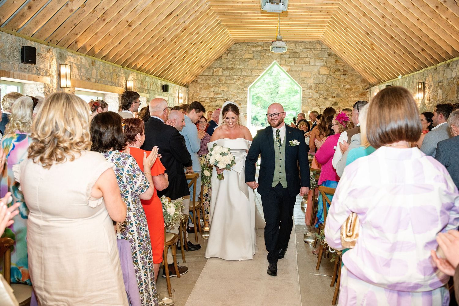 Bride and groom walk down the aisle of a rustic stone chapel surrounded by smiling wedding guests.