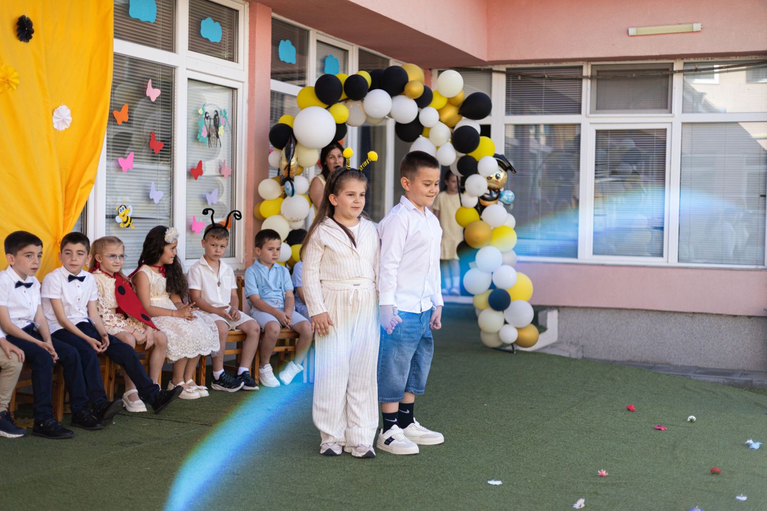 Children performing at a school celebration with balloon arch decor in black, white, and yellow outdoors.