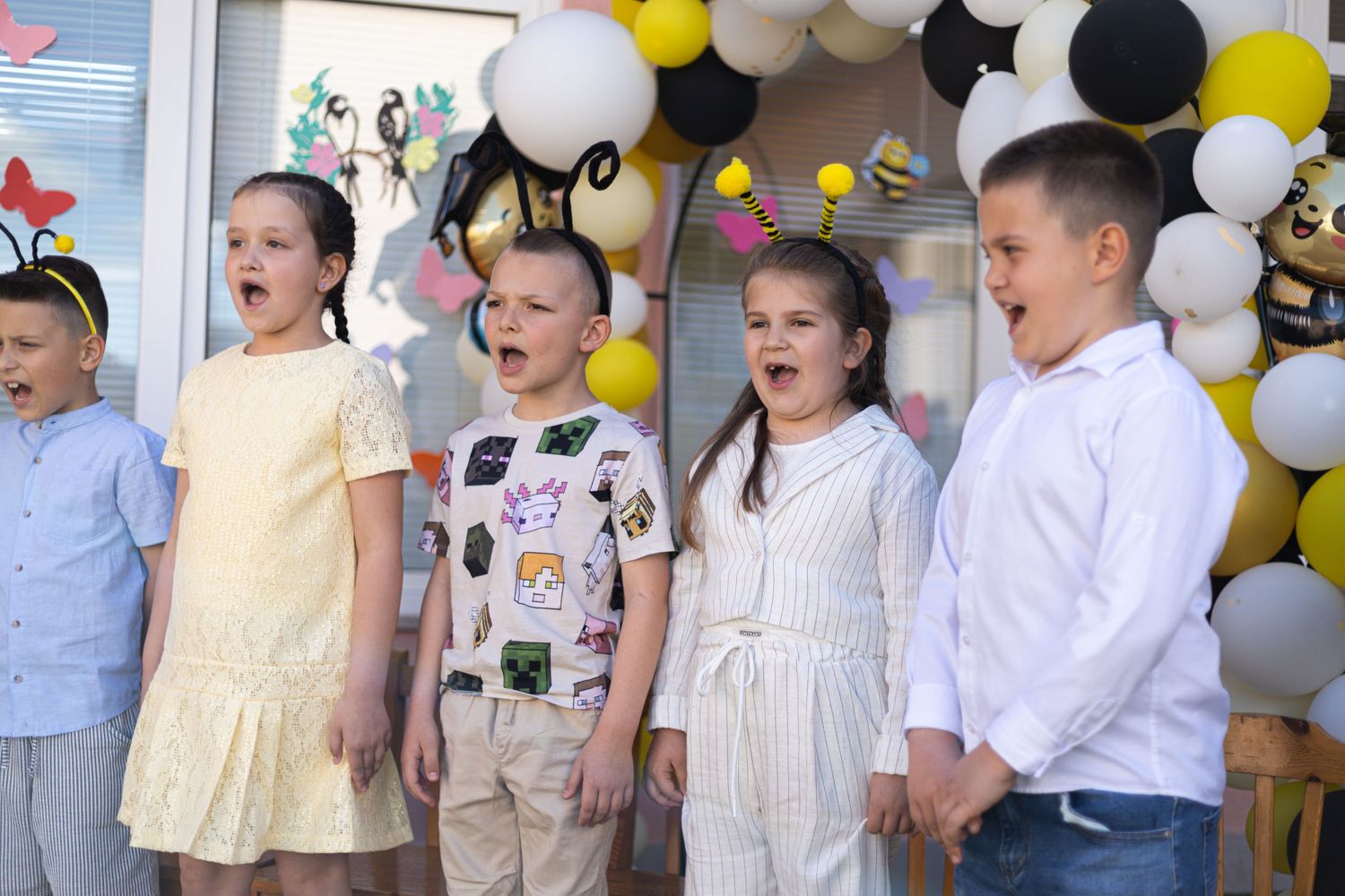 Five children singing at a colorful bee-themed party with black, white, and yellow balloon decorations.