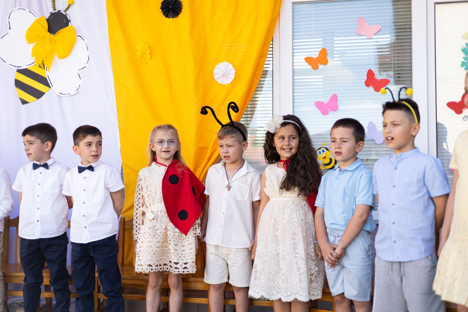 Children in bee and ladybug costumes perform at a school play with colorful decorations.