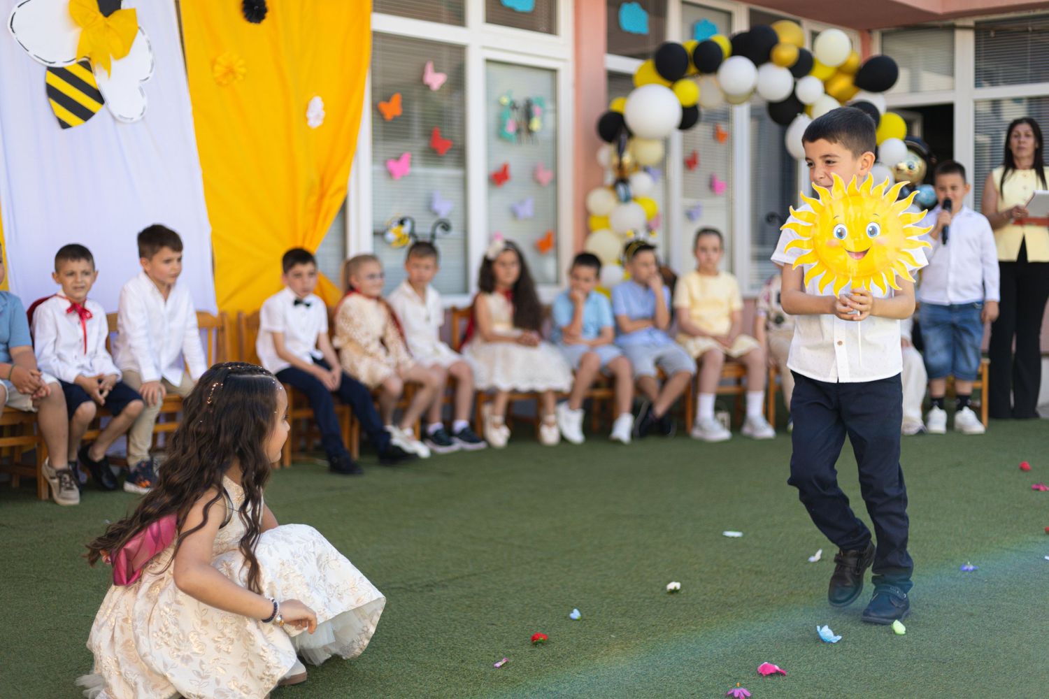 Children performing at a school event with peers watching, colorful bee-themed decorations and balloons in the background.