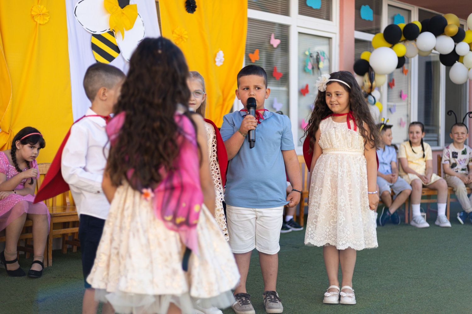 Children performing at a school event outdoors, with a boy speaking into a microphone and a girl in a white dress beside him.