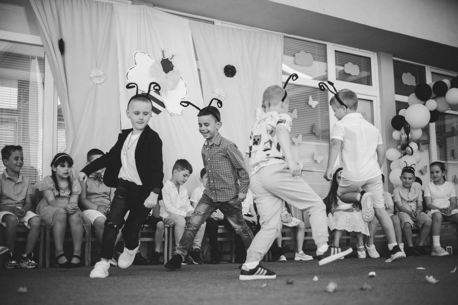 Children in costumes perform a lively dance at a school event, with an audience of parents and kids watching in the background.