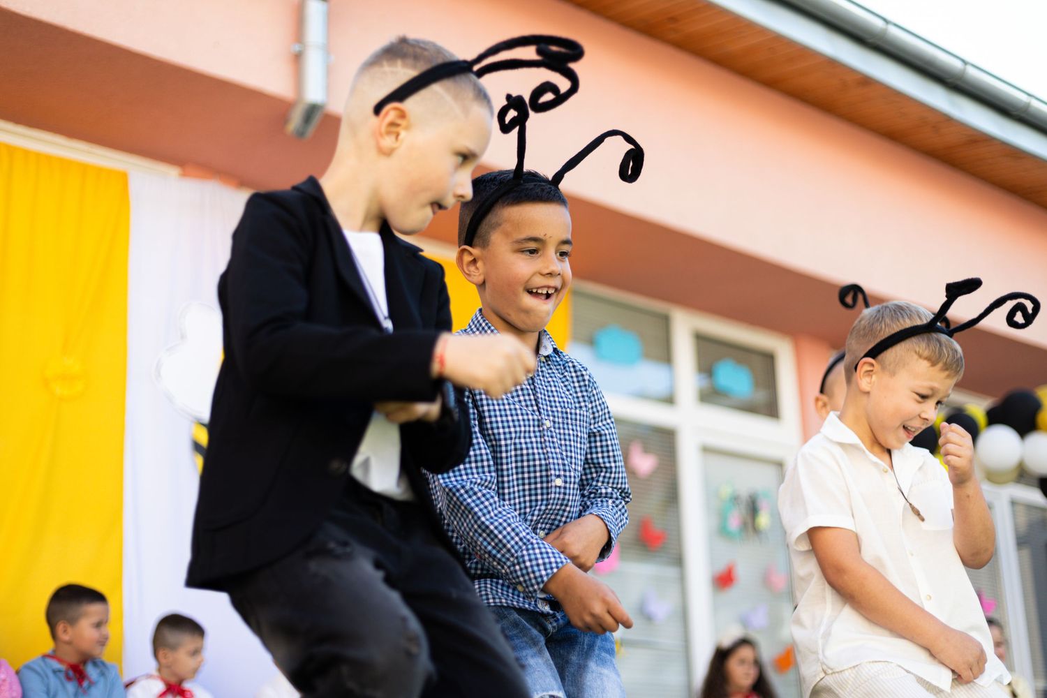 Three children wearing bug antenna headbands laugh and dance together at a school performance outdoors.