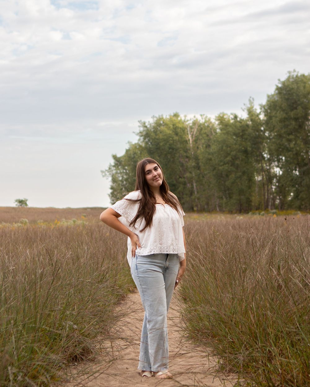 Young woman in white top and jeans smiling on a dirt path through tall grass fields with trees in background.