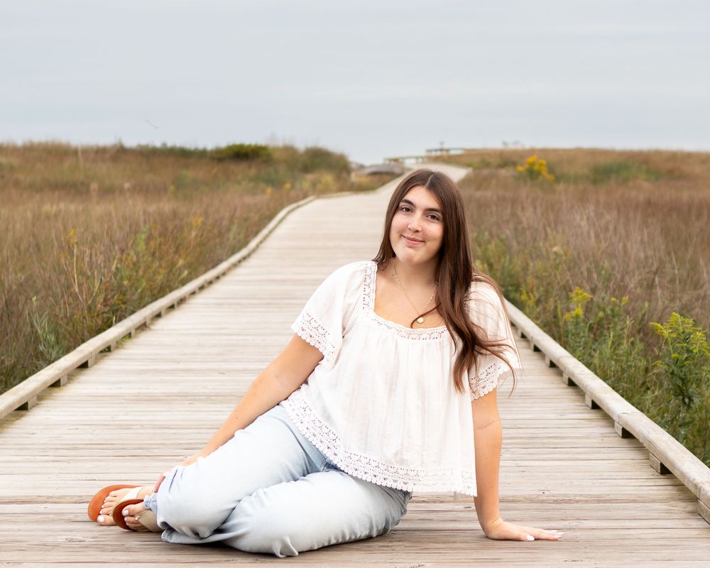 A young woman with long brown hair sits on a wooden boardwalk path through coastal grasses, wearing a white top and jeans.