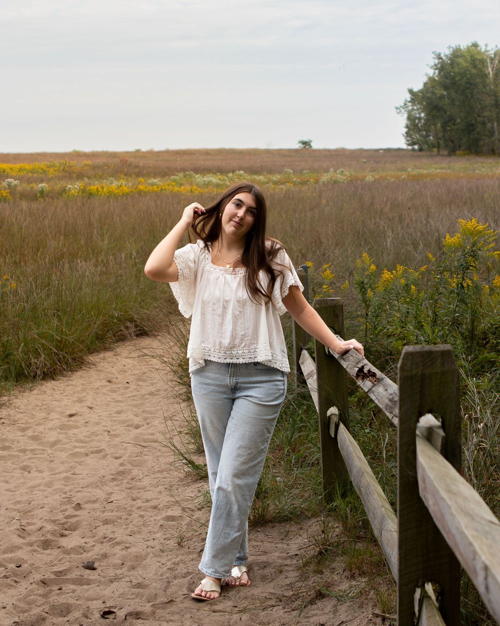Young woman in white top and jeans leaning on wooden fence in a golden meadow path.