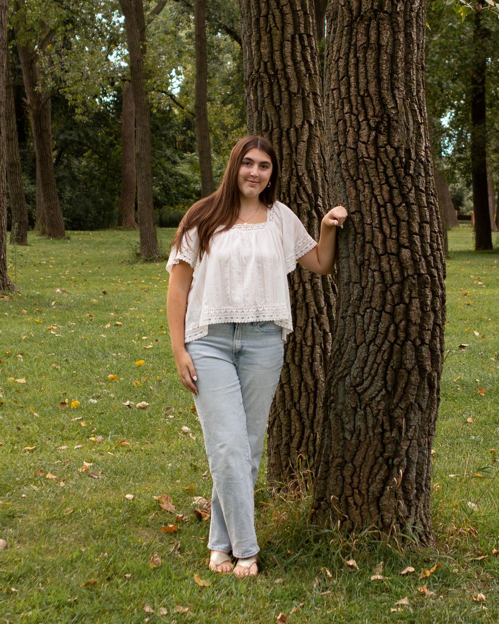 Young woman in white top and jeans leaning against a large tree trunk in a lush green park setting.