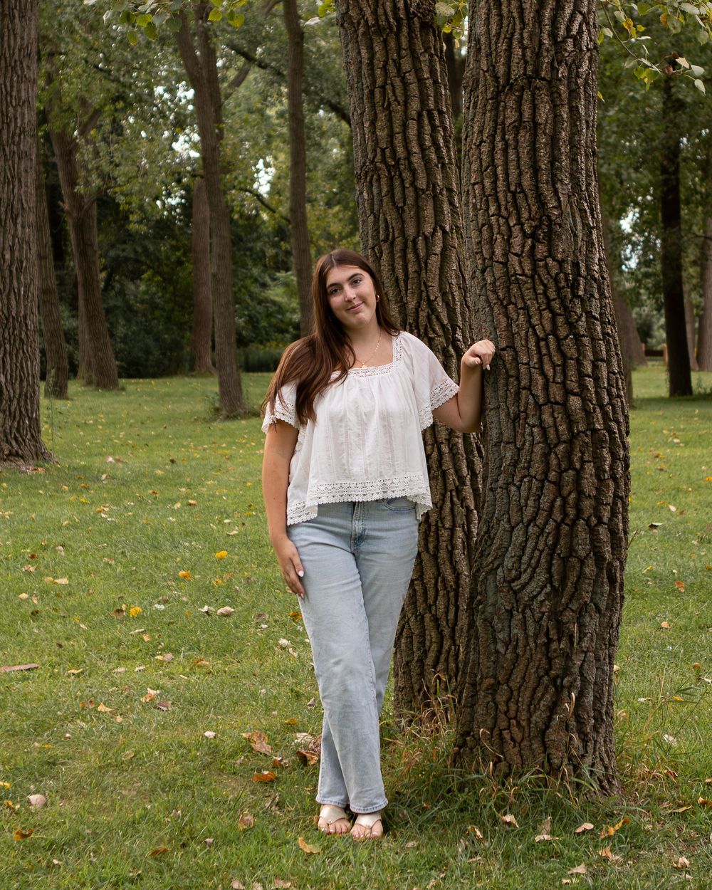 Young woman in white blouse and light jeans posing beside a large tree trunk in a lush green park setting.