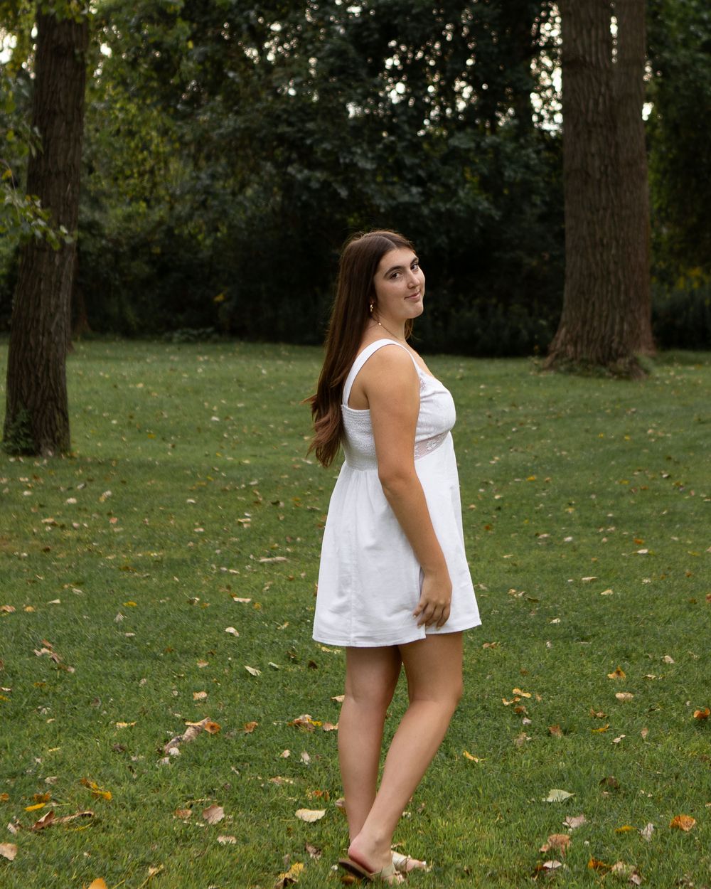 Young woman in white sundress standing on green grass in a park with trees in the background.