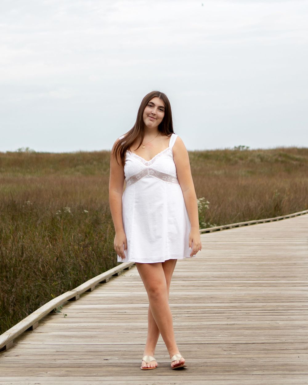 Young woman in white sundress walking on wooden boardwalk path through coastal marsh on cloudy day.