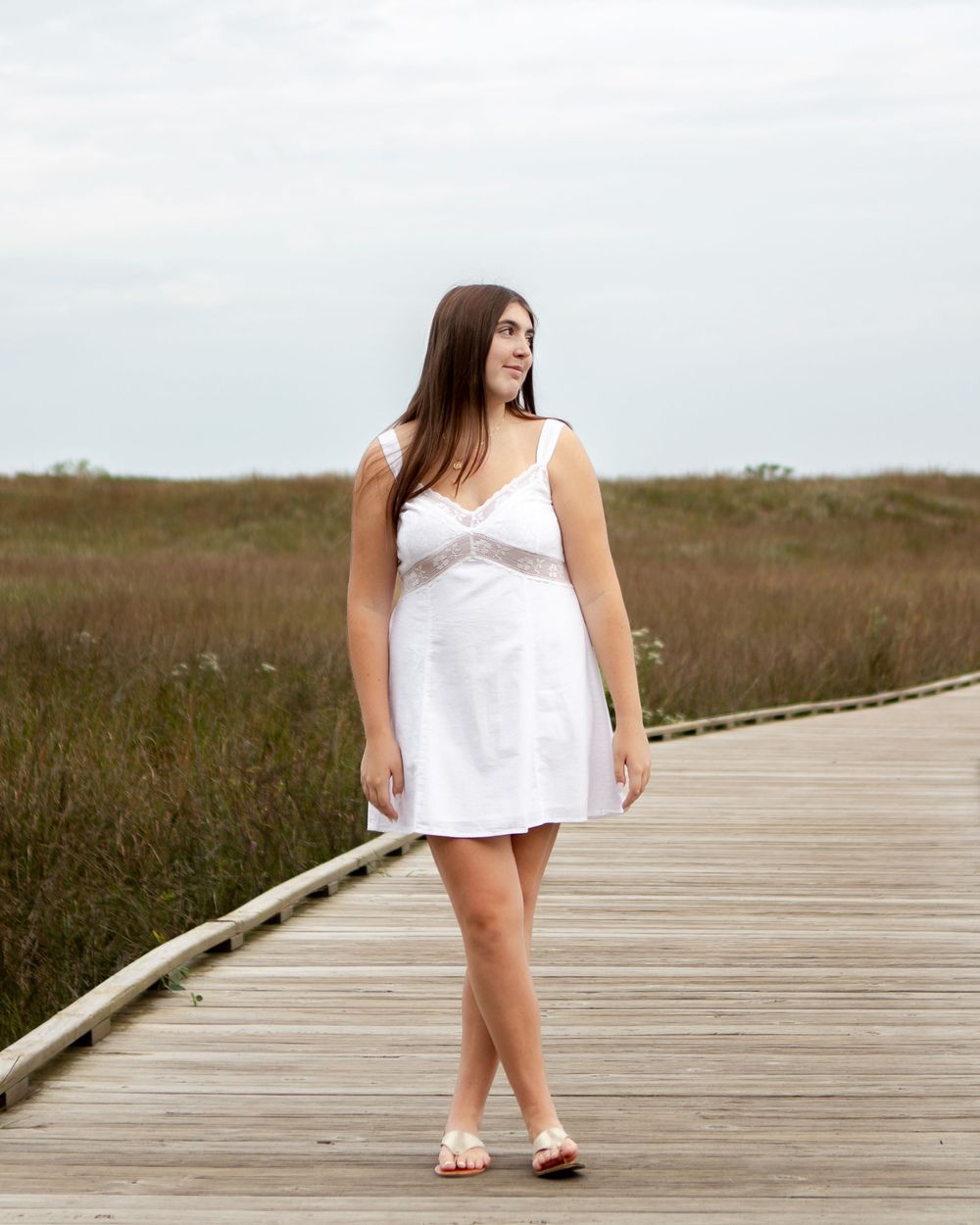 Young woman in white sundress walking on wooden boardwalk through coastal marsh on overcast day.