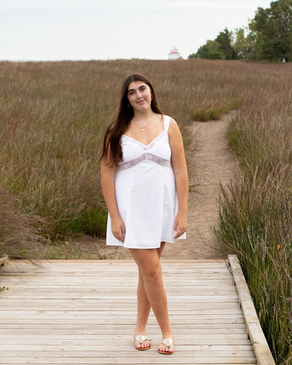 Young woman in white dress standing on wooden boardwalk path through tall grass field near lighthouse.