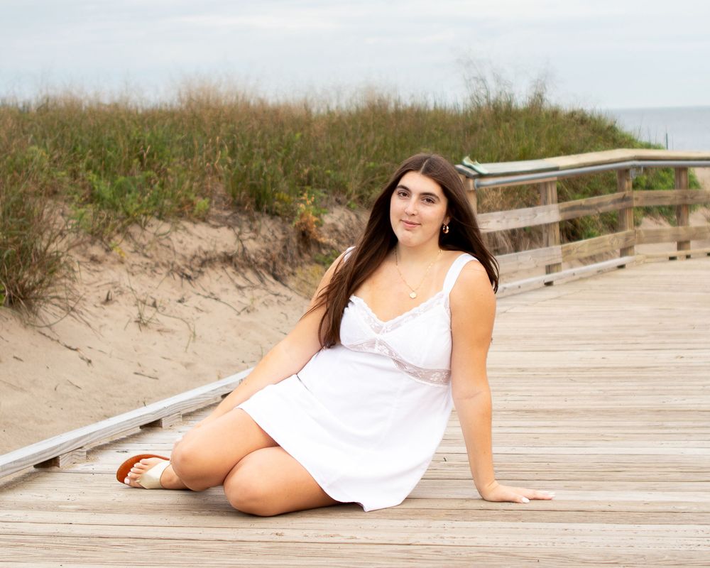 Young woman in white dress sitting on beach boardwalk with sandy dunes behind her.