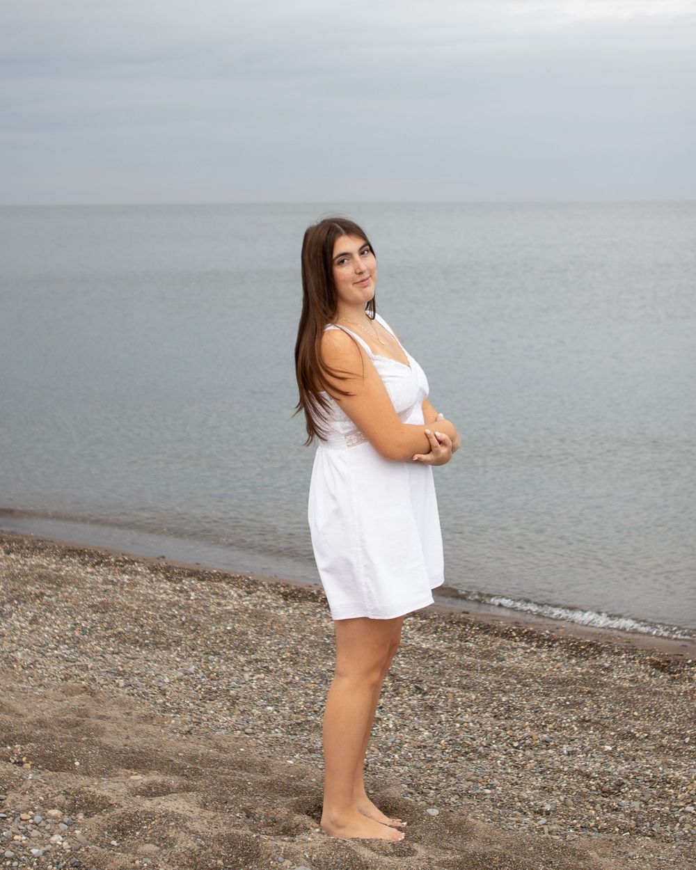 Young woman in white sundress standing barefoot on a pebble beach with calm ocean waters in the background.