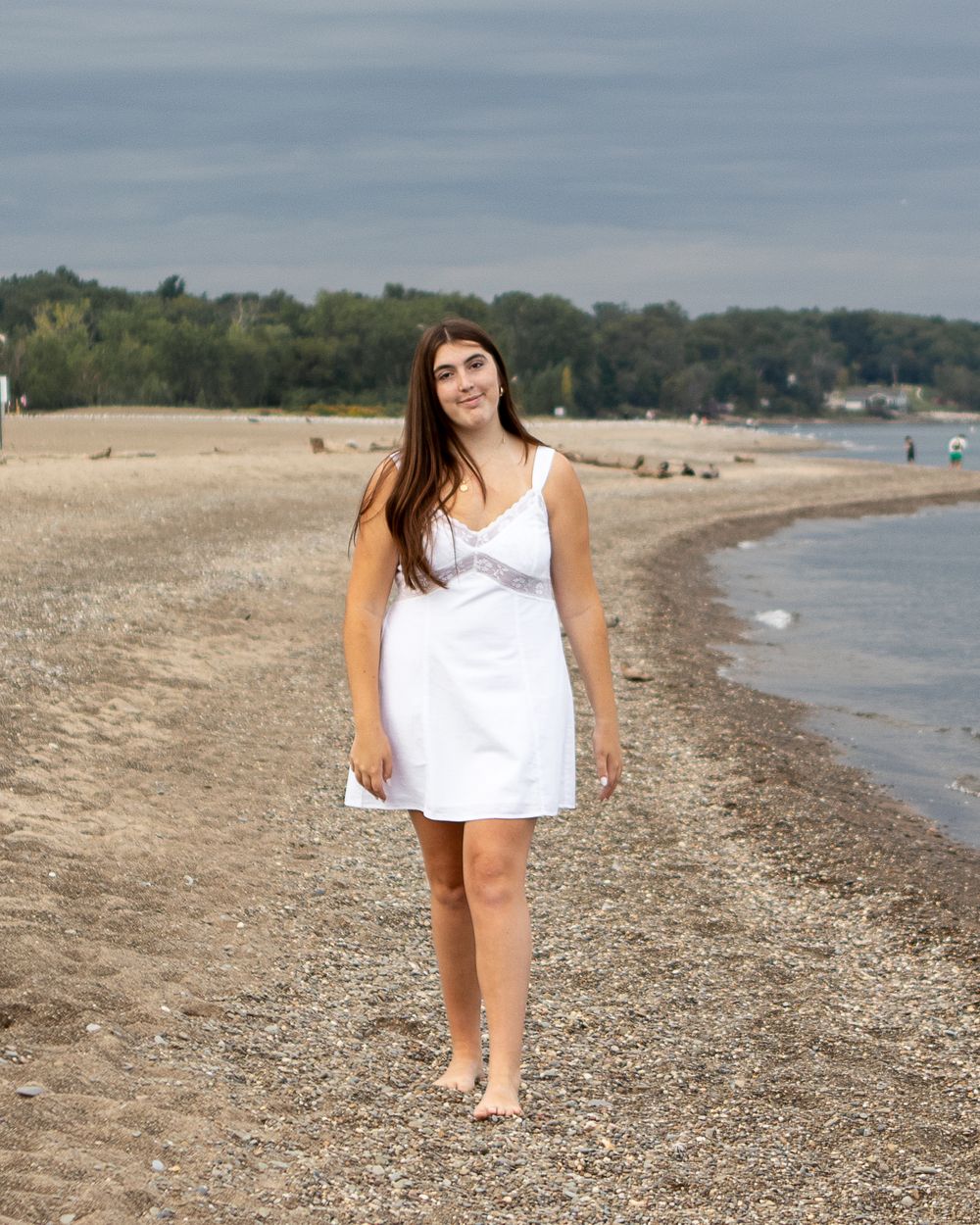 Young woman in white sundress standing barefoot on a rocky beach with trees and cloudy sky in background.