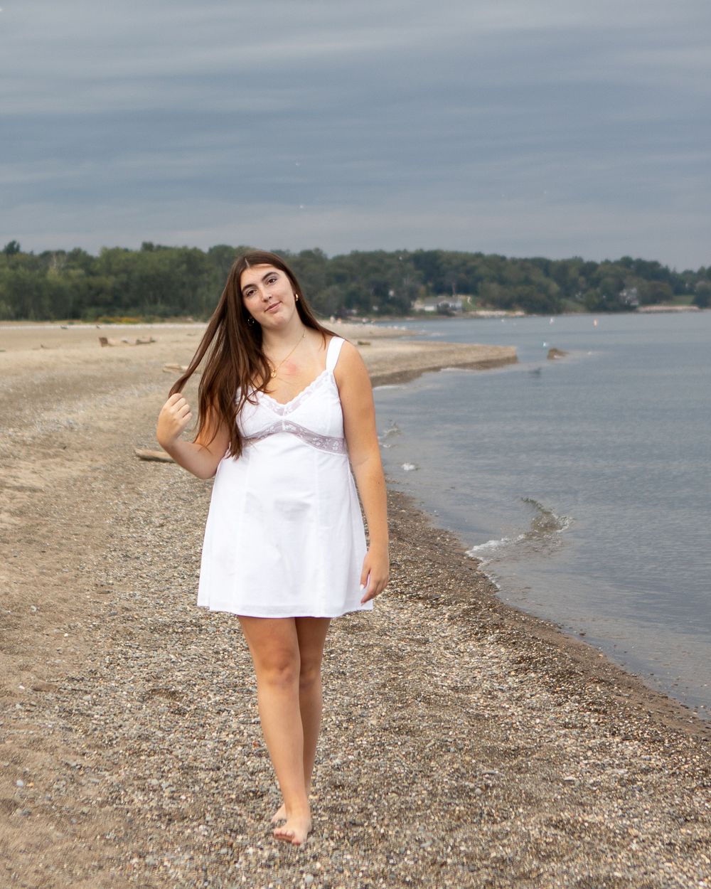 Young woman in white dress walking barefoot on a rocky beach with calm water and trees in the background.