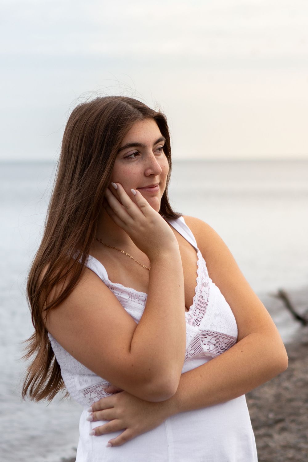 Young woman in white lace dress poses thoughtfully by the ocean, hand on cheek, long brown hair flowing.