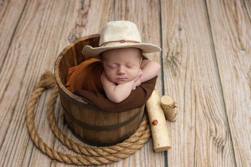 Newborn baby sleeping in wooden bucket wearing cowboy hat with rope and logs on wood floor.