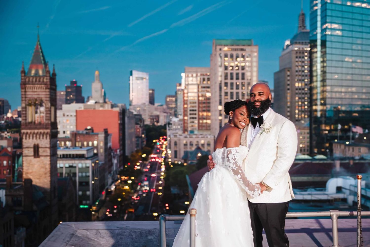 Bride and groom posing together on the rooftop deck of the Lenox Hotel in Boston at dusk with the skyline behind them.