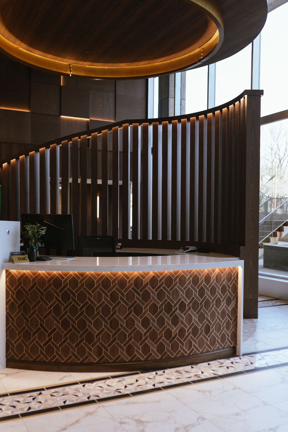 Elegant hotel lobby with a geometric reception desk, curved staircase, marble floors, and warm ambient lighting.