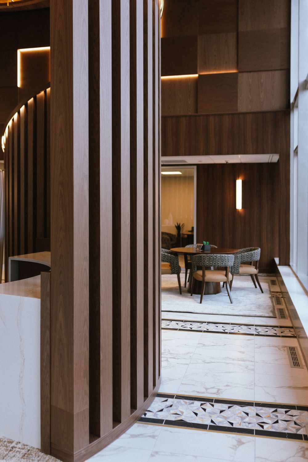 Modern hotel lobby with marble floors, wood panel walls, pendant lighting, and elegant dining area with chairs and tables.