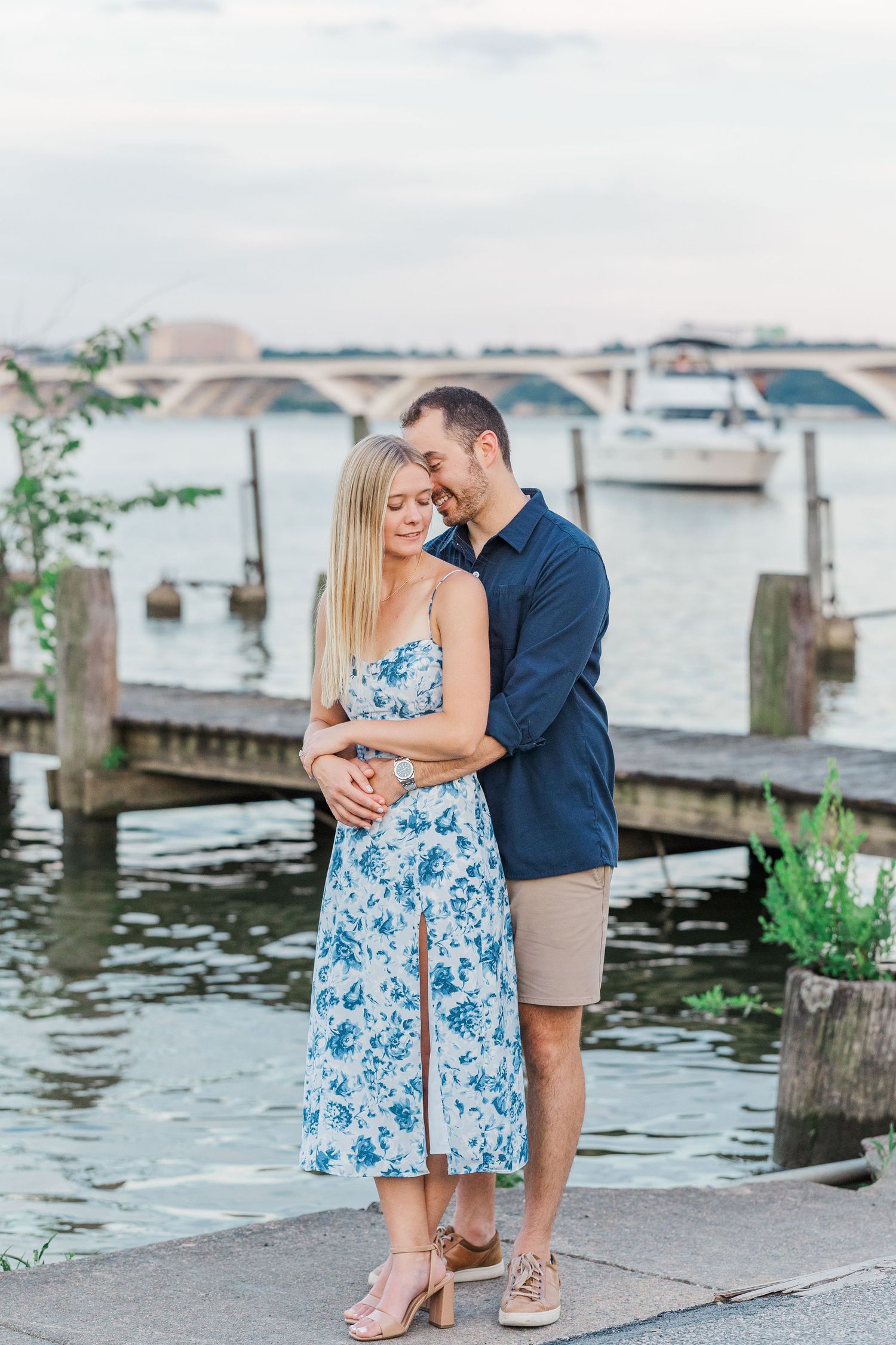 Couple embracing on a waterfront dock, woman in floral dress and man in navy shirt, with a bridge and boats in background.