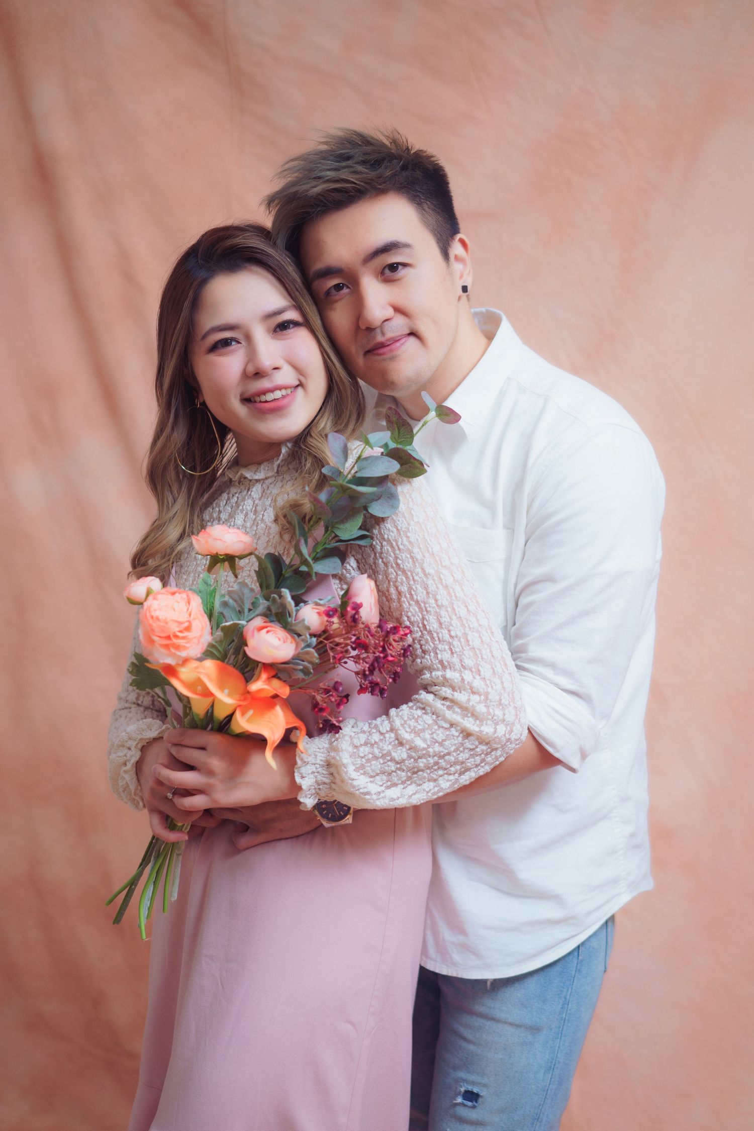 A couple poses against a peach backdrop while holding a vibrant bouquet of coral roses and eucalyptus.