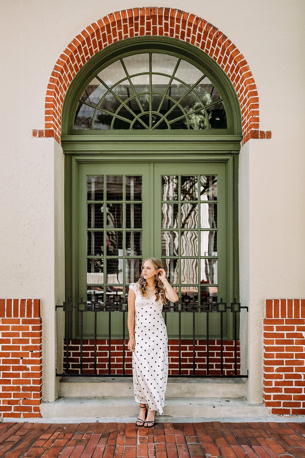 Woman in white polka dot dress posing before elegant green arched double doors with brick accents.