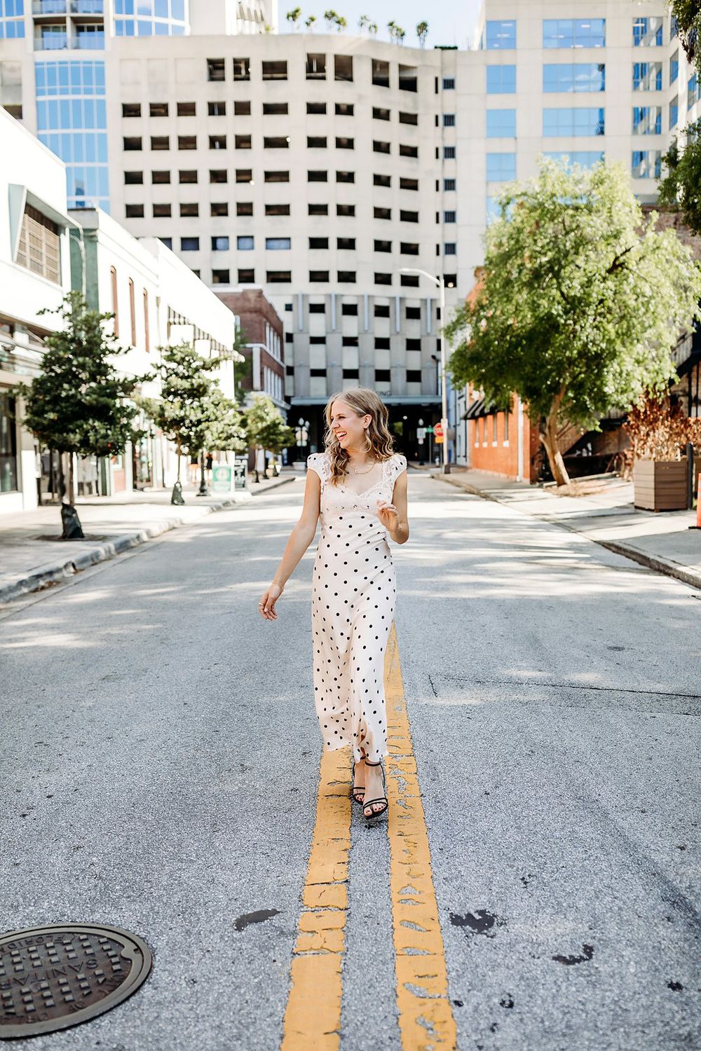 Woman in white polka dot maxi dress walking on urban street centerline with city buildings and trees in background.