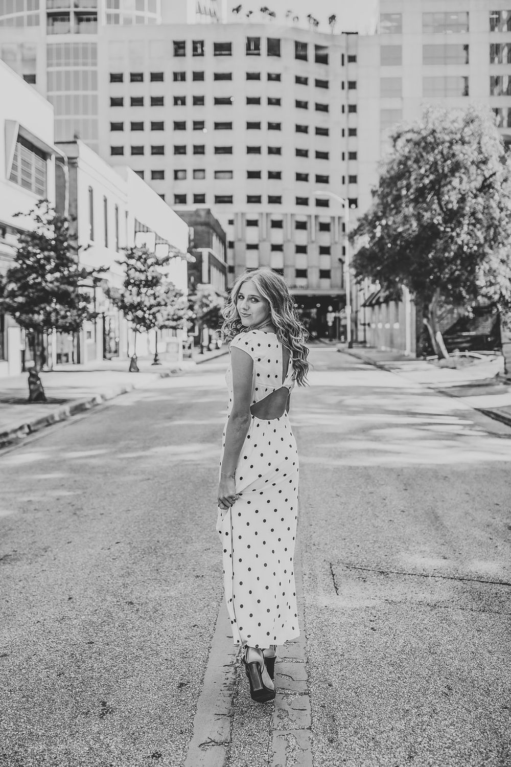 Black and white photo of a woman in a polka dot maxi dress posing on an urban street with city buildings.