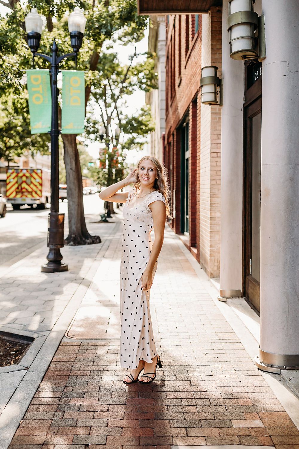 Blonde woman in white polka dot maxi dress posing on a brick sidewalk in a sunny urban downtown setting.