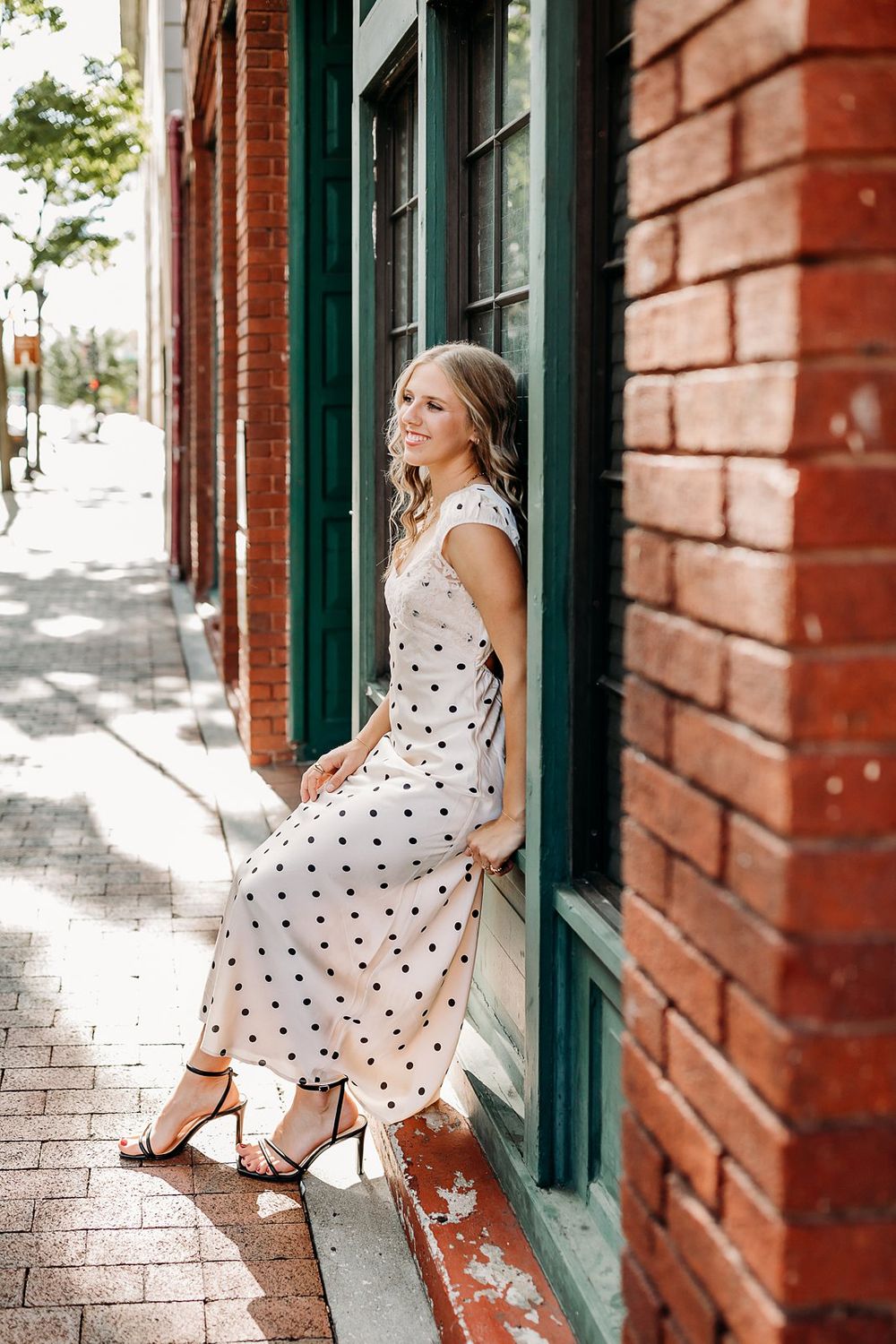 Young woman in white polka dot midi dress and heeled sandals posing against brick building on sunny day.