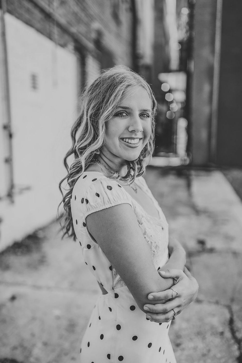 Black and white portrait of a smiling young woman in a polka dot dress, posing outdoors in an urban alley.