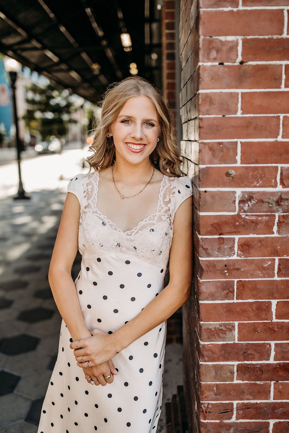 Young blonde woman in polka dot dress smiling against brick wall outdoors.
