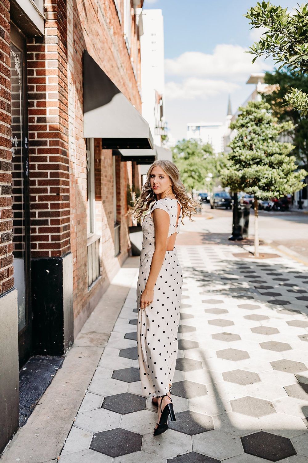 Woman in polka dot maxi dress and black heels posing on a hexagonal tile sidewalk beside a brick building.