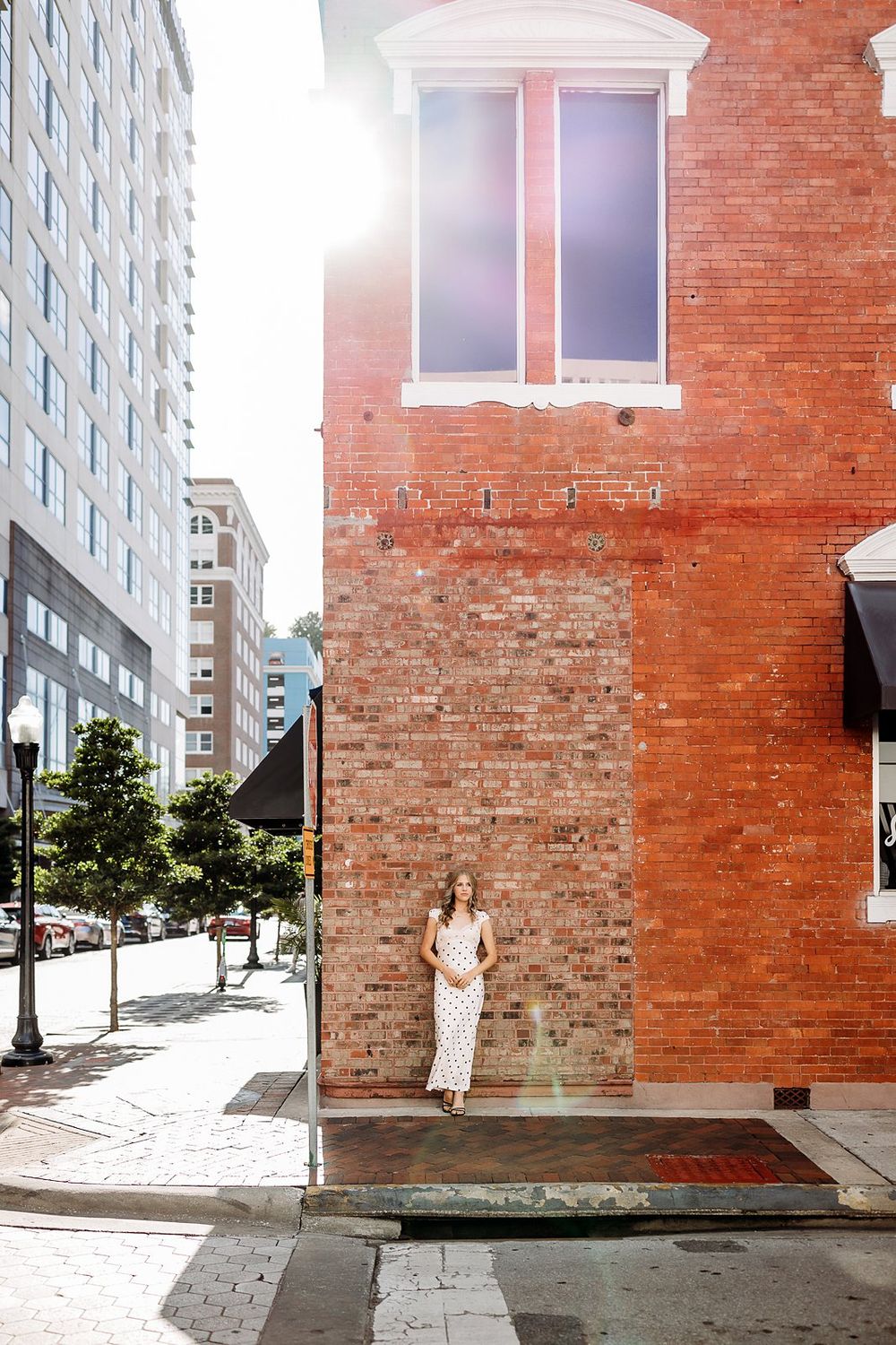 Woman in polka dot midi dress standing against red brick building corner in sunny urban setting.