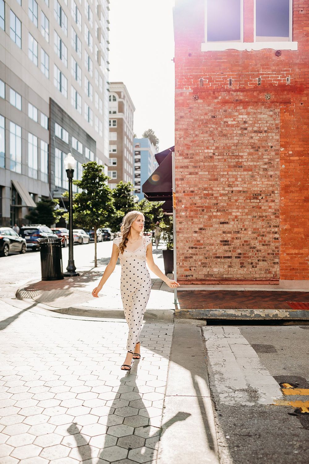 Woman in white polka dot dress walking on city sidewalk beside red brick building in sunlight.