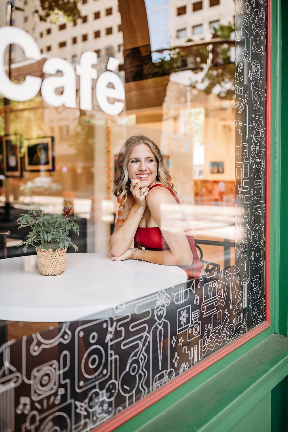 Smiling blonde woman in red top sitting at a white cafe table, viewed through a decorated window.