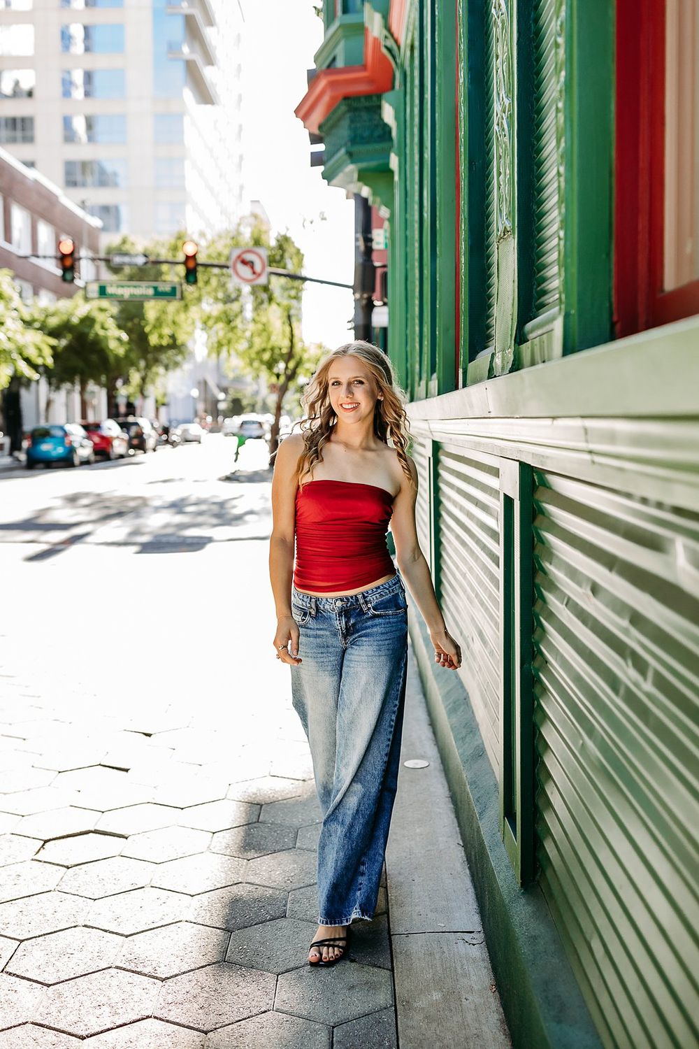 Blonde woman in red tube top and wide-leg jeans leaning against green building on sunny urban sidewalk.