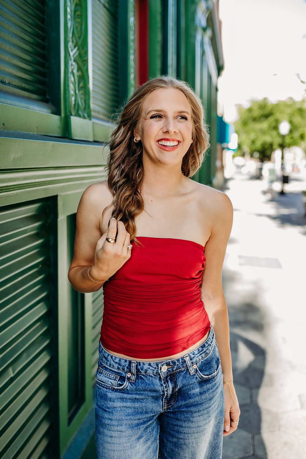 Smiling young woman in red tube top and jeans posing outdoors near green building on sunny day.