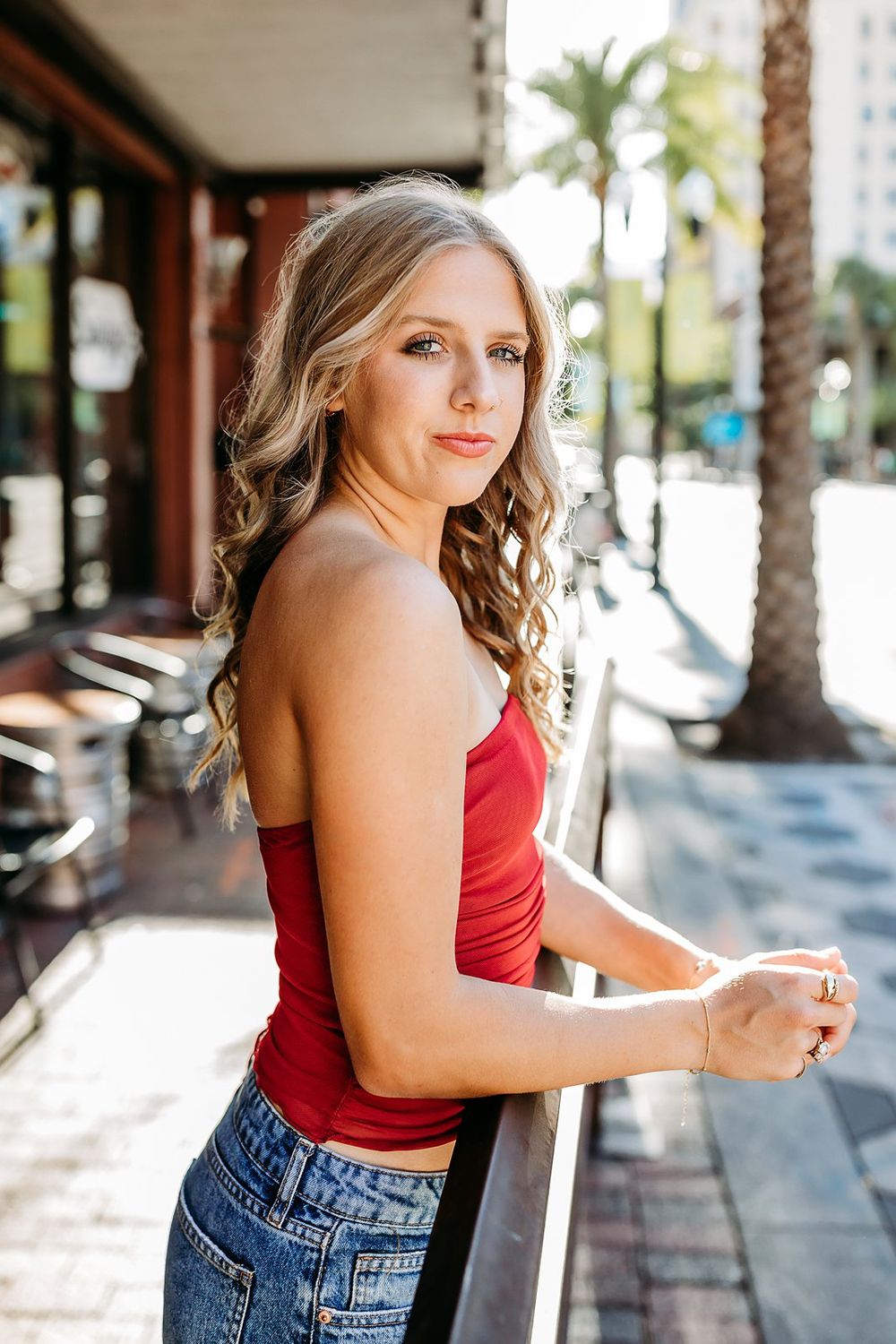 Young woman in red crop top and jeans leaning on railing with palm trees on sunny urban street.