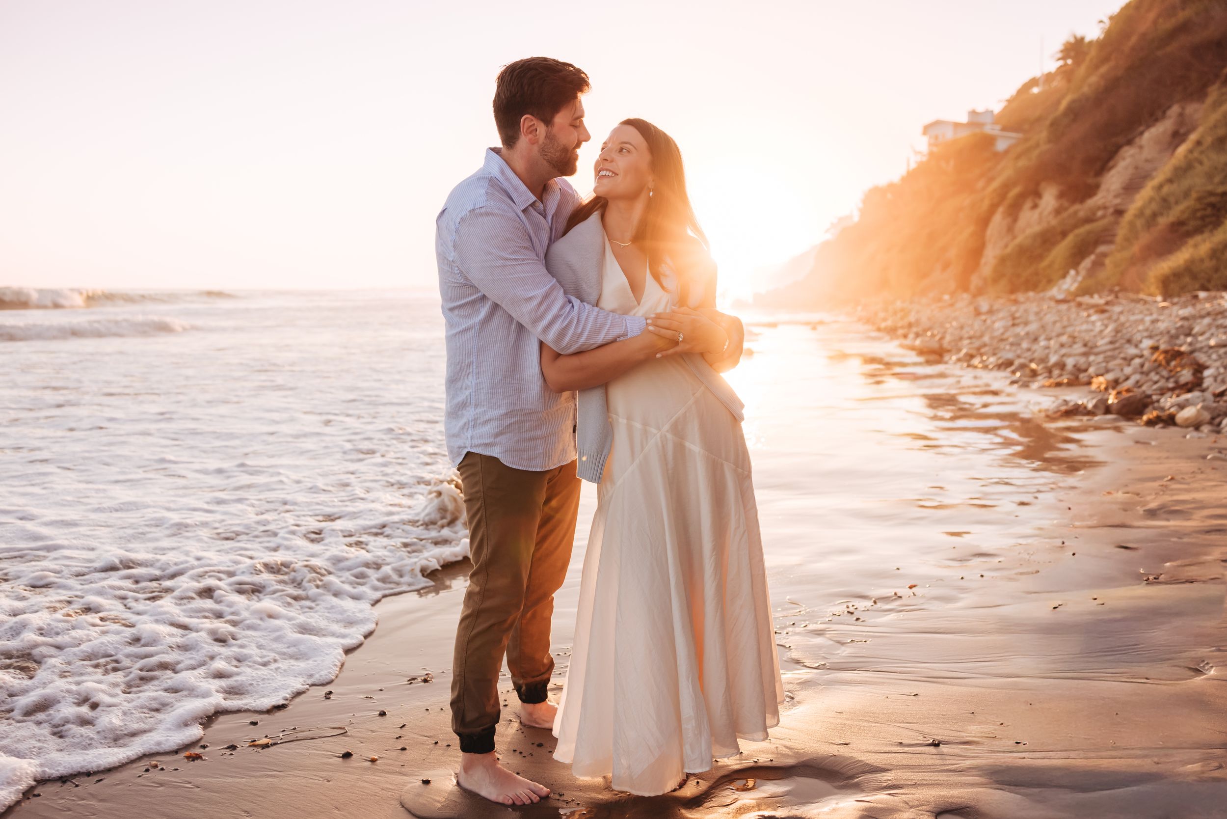 Newley engaged couple wearing white smiling and toasting champagne while sitting on a beach in Santa Barbara