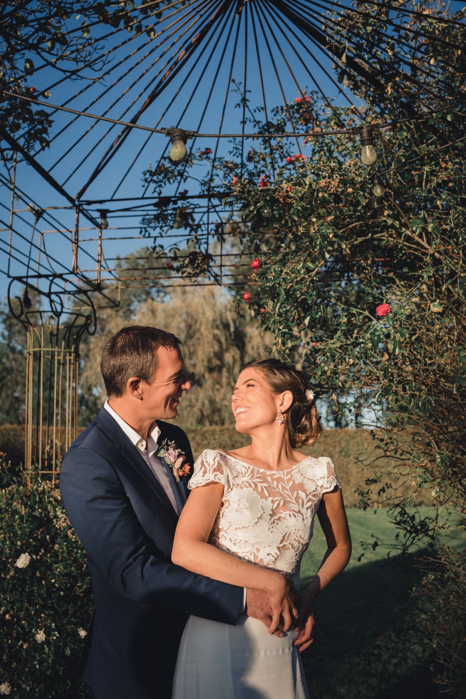A smiling bride and groom embrace outdoors under a decorative iron arbor with roses in warm golden light.